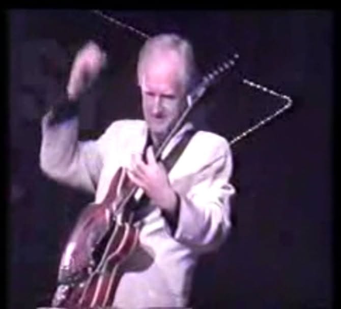 Allen in a pale jacket playing his red Gibson on a dark stage, one hand raised at the end of a strum